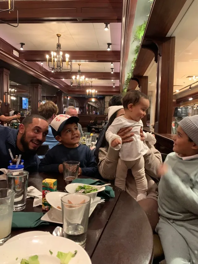 The Family is gathered around the dinner table at a restaurant. There is a man, a woman, and three small children. They are all facing the small little girl who is held by the woman.