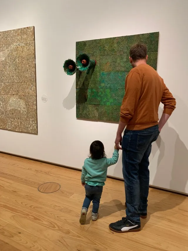 Small child and father admiring an art installation hanging on a wall that appears to look like a painting from afar.