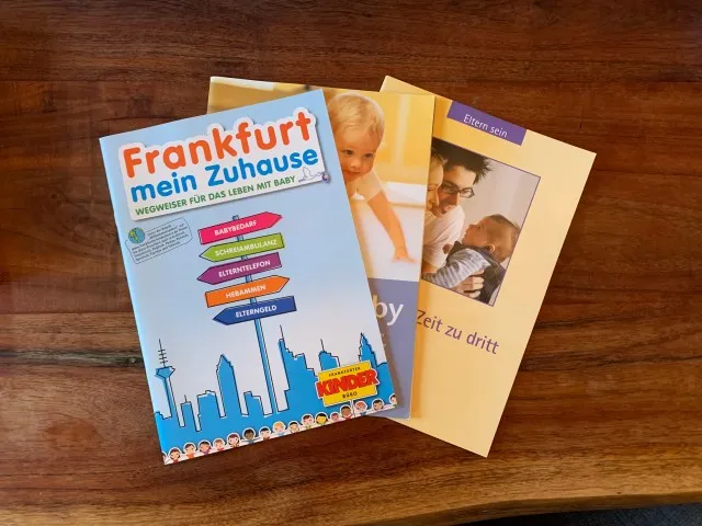 Three different Pamphlets are on top of a wooden dining table which include information on Frankfurt and life with a baby and as new parents. All three pamphlets look like informational magazines.