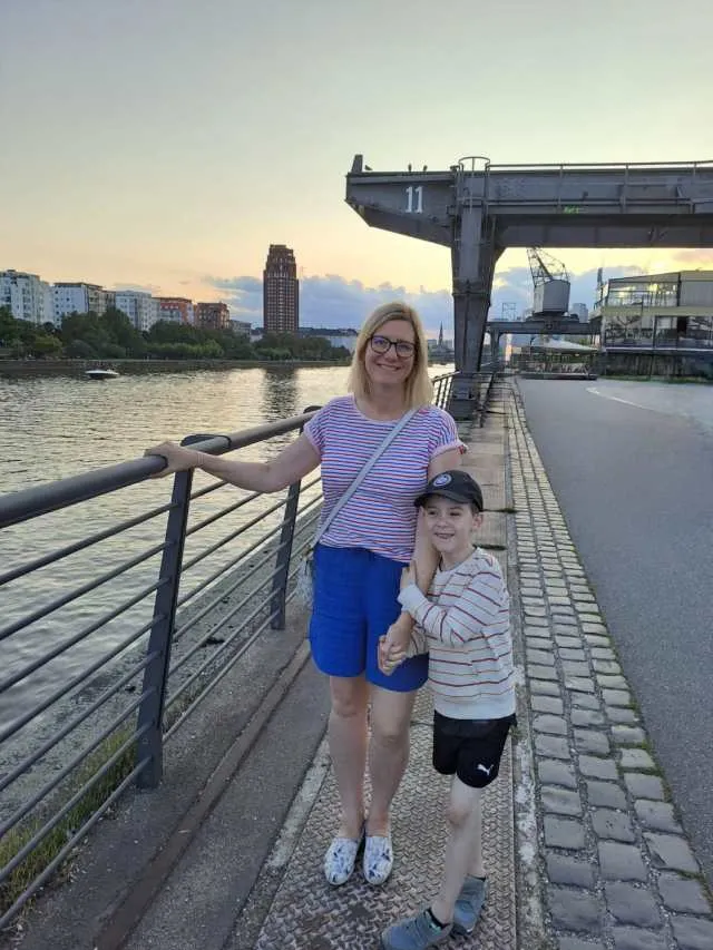 Mother and son stand next to each other while the son holds the mother's arm. It is summertime and they have a view of Frankfurt in the background with the Main River.