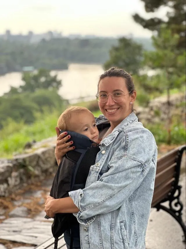 A woman holding her baby in a baby carrier. The background has a lake view.